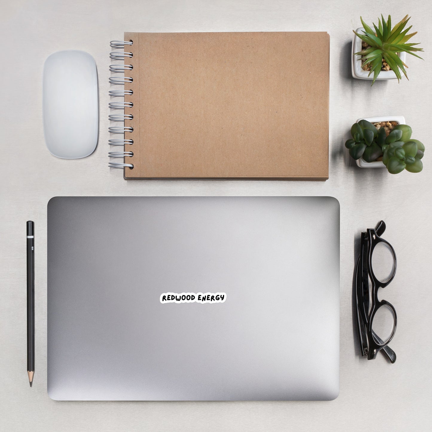 Laptop with 'Redwood Energy' sticker on a desk with notebook, mouse, pen, and plants.
