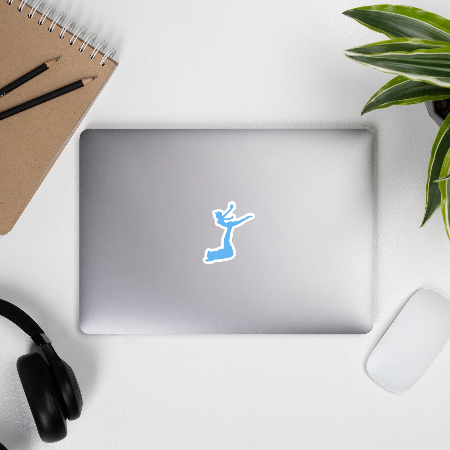 Air Jordon Logo of a man holding a woman in blue stuck to a laptop with two pencils on a notebook, the side of a headphone, a mouse, and the leaves of an office desk plant surrounding the laptop.